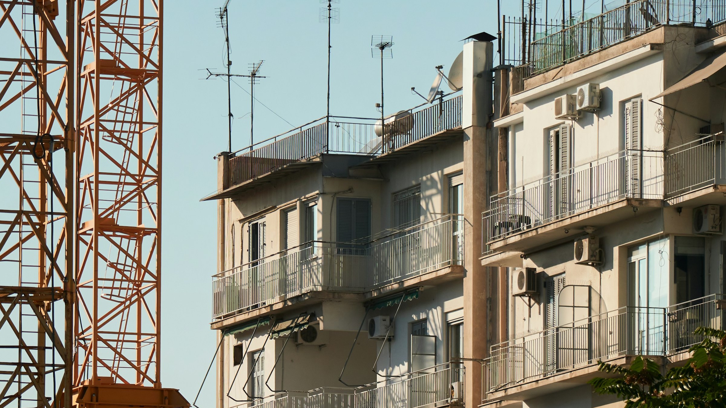 A typical Athens polykatoikia, a multi-storey apartment block of flats. Roof antennas, satellite dishes and air-conditioning units bolted to the balconies. A construction crane in the foreground. Documentary photograph of the kind of stock that holds the long tail of Greek residential tenancy.