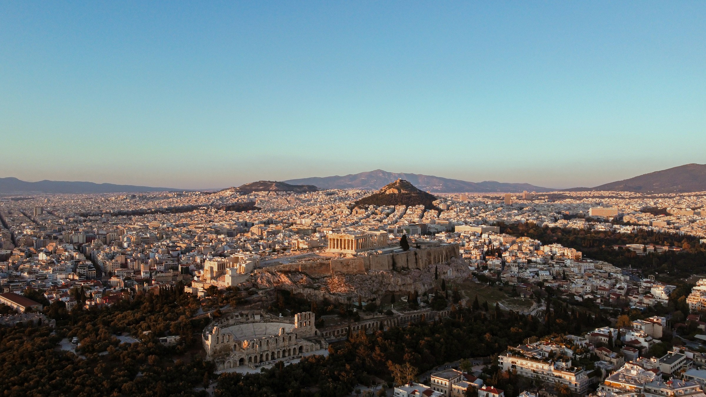 An aerial photograph of dense Athens residential blocks, the close-packed rooftops of central neighbourhoods that the Golden Visa programme has transacted across, from inside the ring road outward to the suburbs.