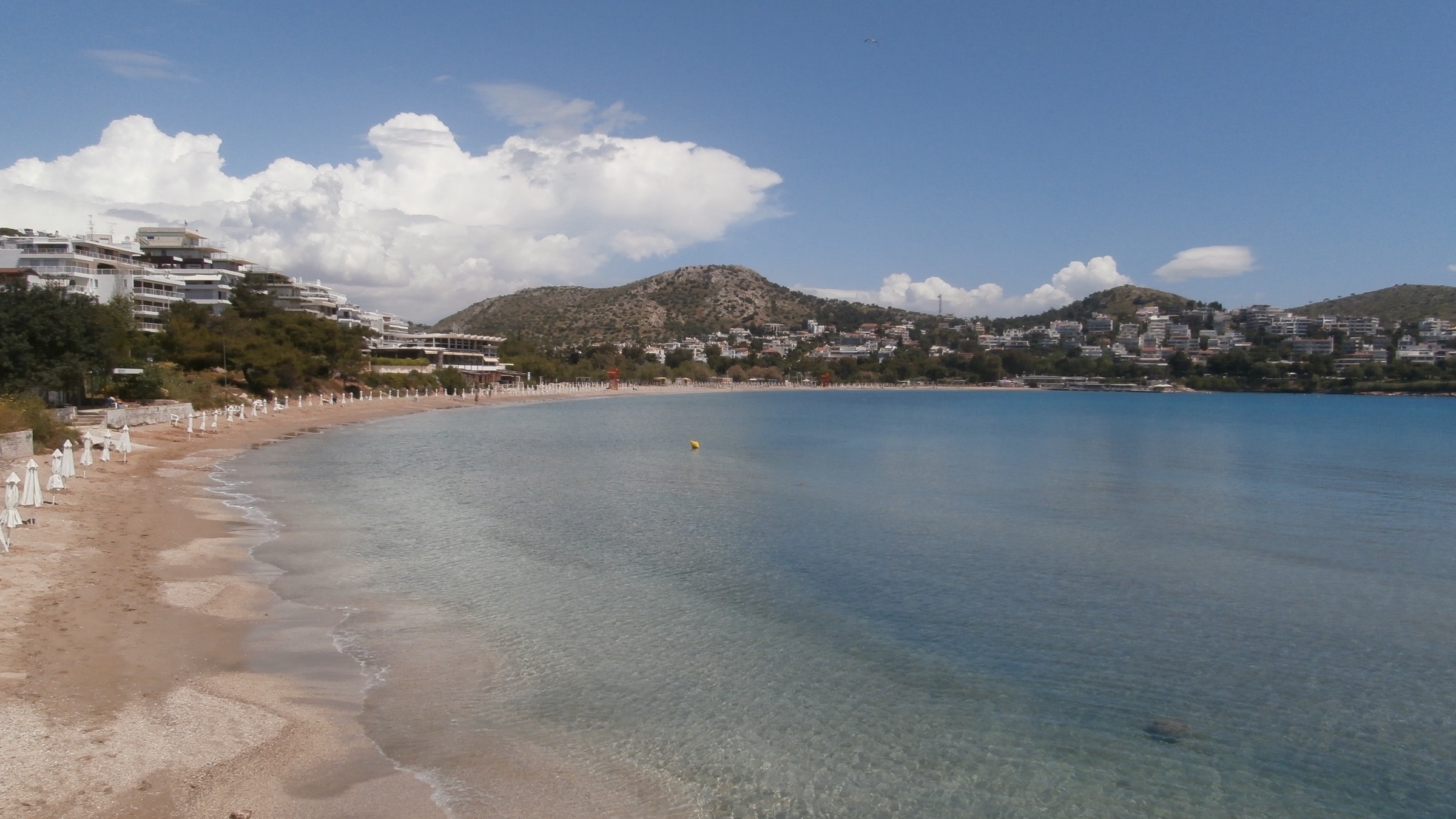 The Athens Riviera coastline at Vouliagmeni: a turquoise bay ringed by hillside residences and pine headlands. The stretch of coast where Greece's branded-residence pipeline is concentrated.