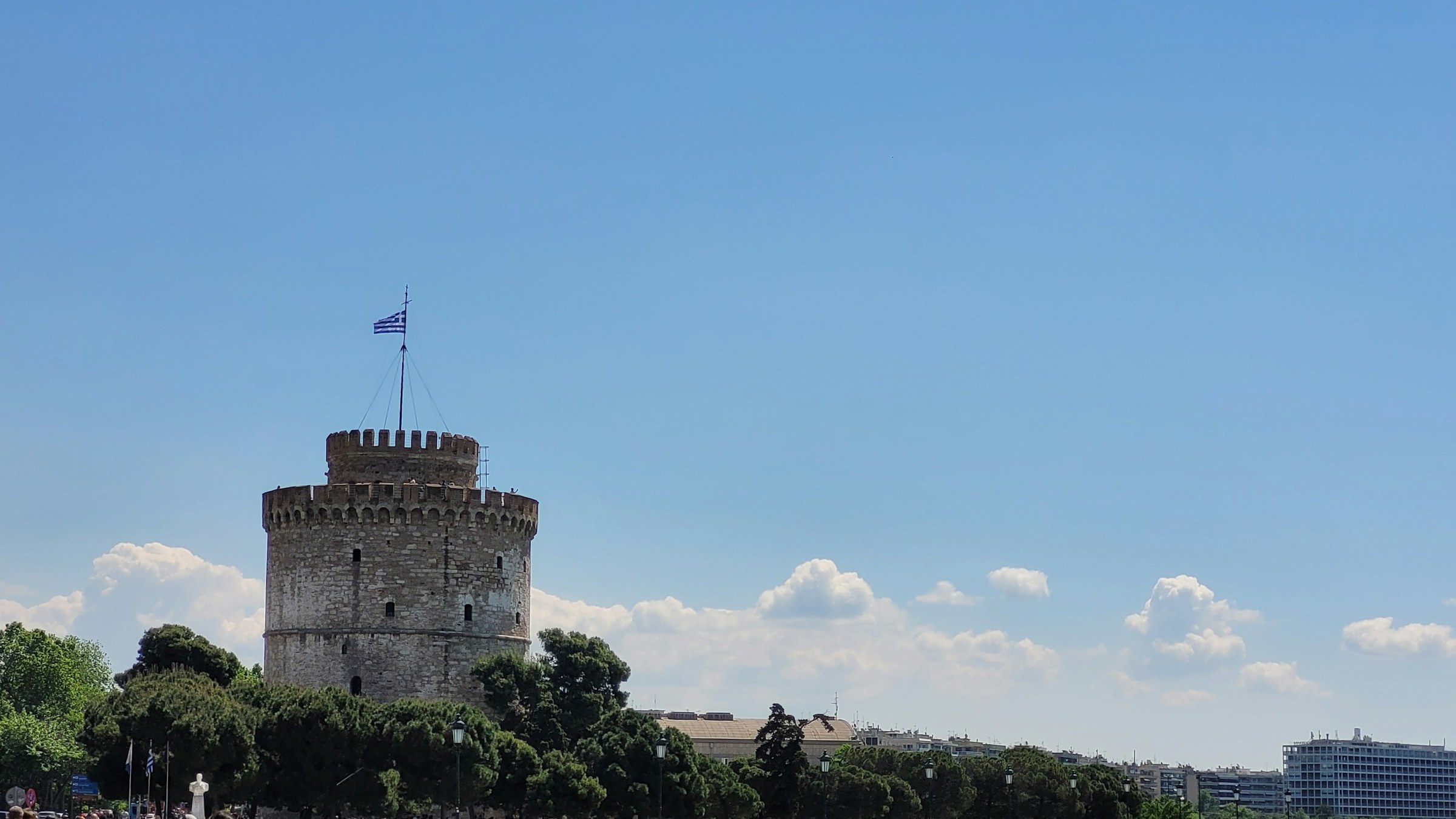 The White Tower of Thessaloniki on the city's waterfront under a clear sky. The cylindrical 15th-century landmark anchors the harbour edge that has framed every redevelopment cycle the city has been through.