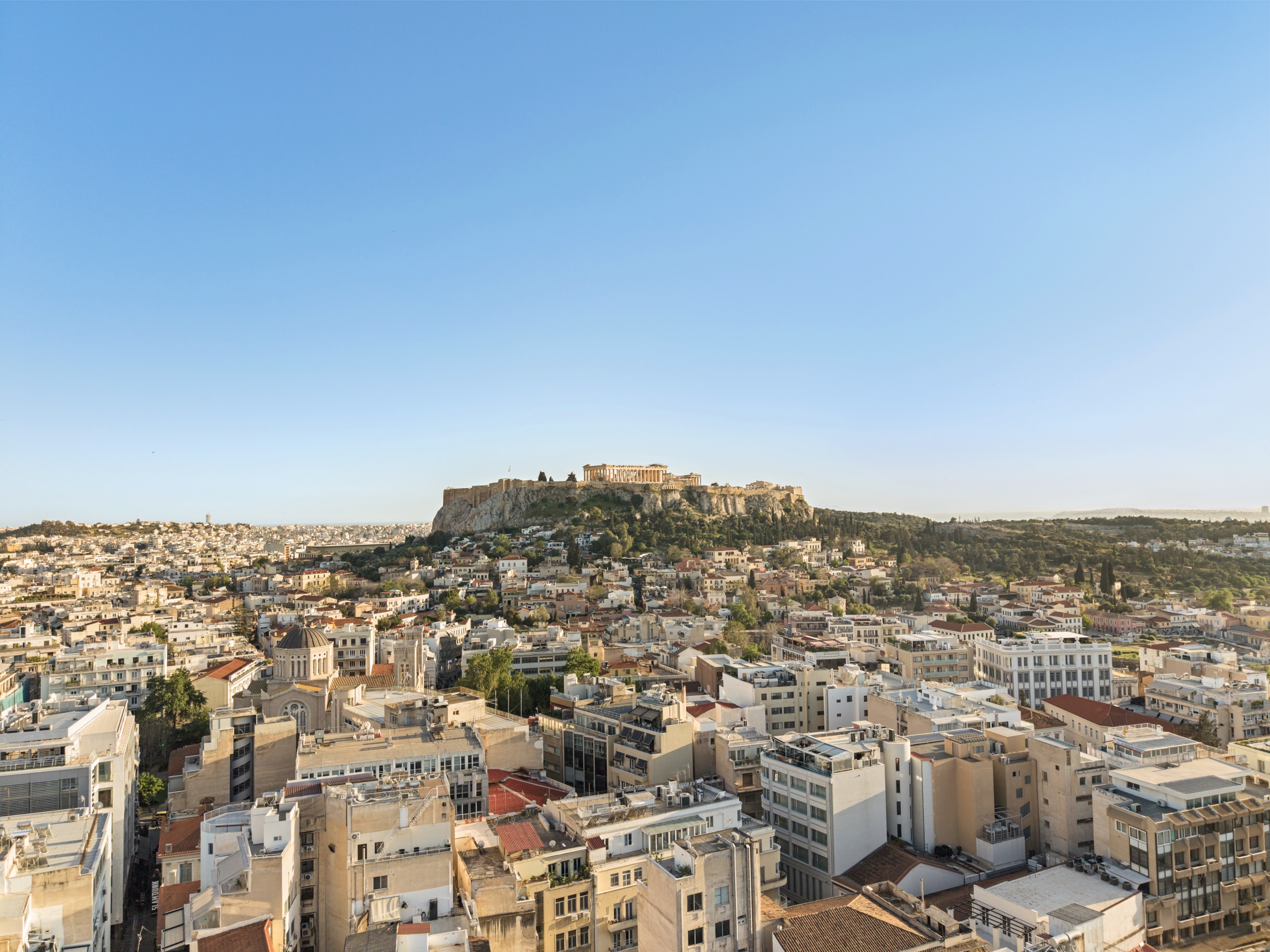 The Acropolis of Athens rises above the low-rise city, viewed from a rooftop in central Athens at golden hour. The Parthenon is clearly visible on the rock.
